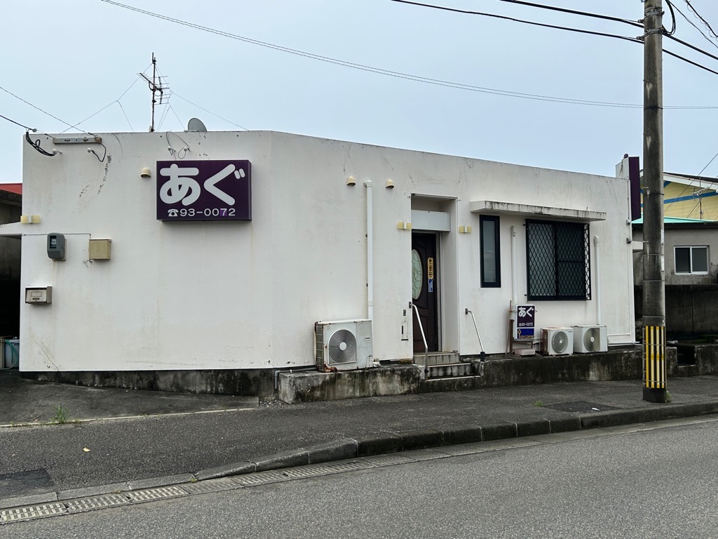 白い平屋建ての建物を正面から捉えた写真で、壁には、「あぐ」と電話番号が書かれた紫色の看板が正面と玄関脇にある物件の外観写真