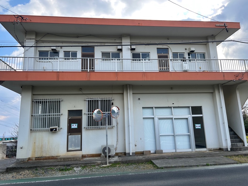 白とオレンジ色で塗り分けられた2階建ての建物が真正面から捉えられ、1階には格子状の窓とガラス張りの店舗入口があり右側には2階に上がる階段があるアパートの外観写真