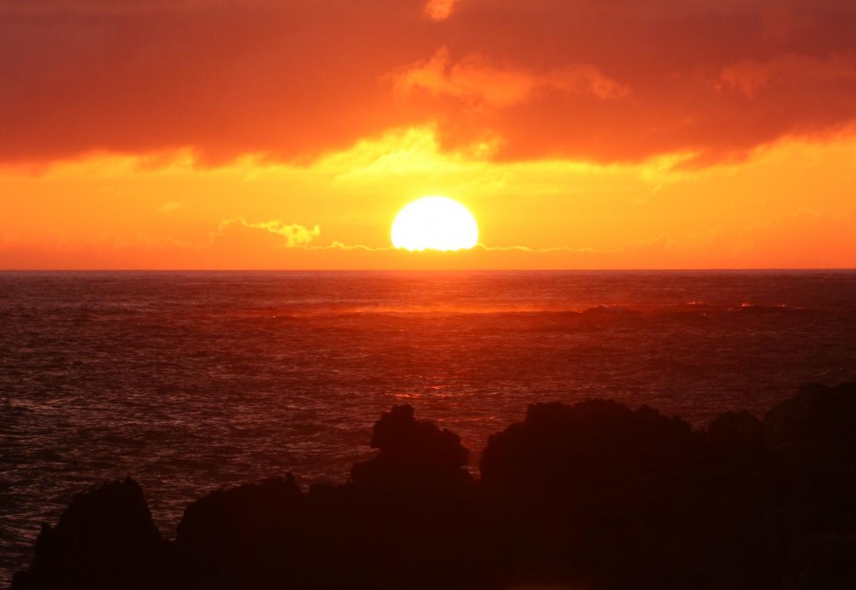 燃えるようなオレンジ色と黄色に染まった空の低い位置に、水平線上の海を照らしながら、まばゆい半円形の太陽が沈む夕焼けの写真