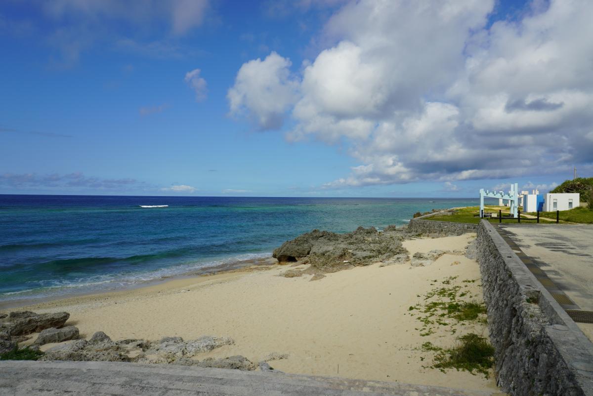 晴れ渡る青空の下に広がる透明度の高いエメラルドグリーンの海と白い砂浜、その傍らには石垣の防波堤と遊歩道が整備され、遠くに白い建物や東屋が見える穏やかな屋子母海岸の風景写真