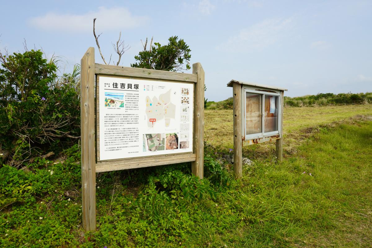 緑の草地に囲まれた住吉貝塚について、場所や説明、地図などが記載されている木製の大きな案内板と、右奥にあるガラスケース付きの小さな掲示板を写した写真