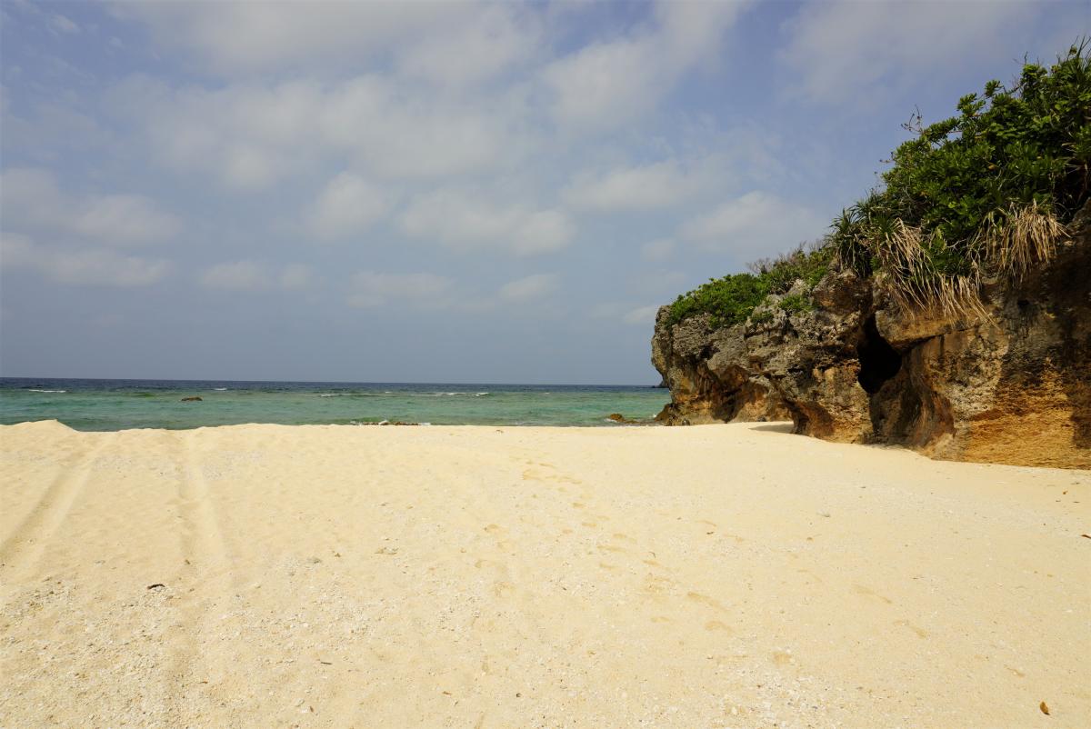 白い砂浜が広がり、右側には緑の植物に覆われた茶色い岩の崖が海に突き出しており、青い海が広がる友留の浜の写真