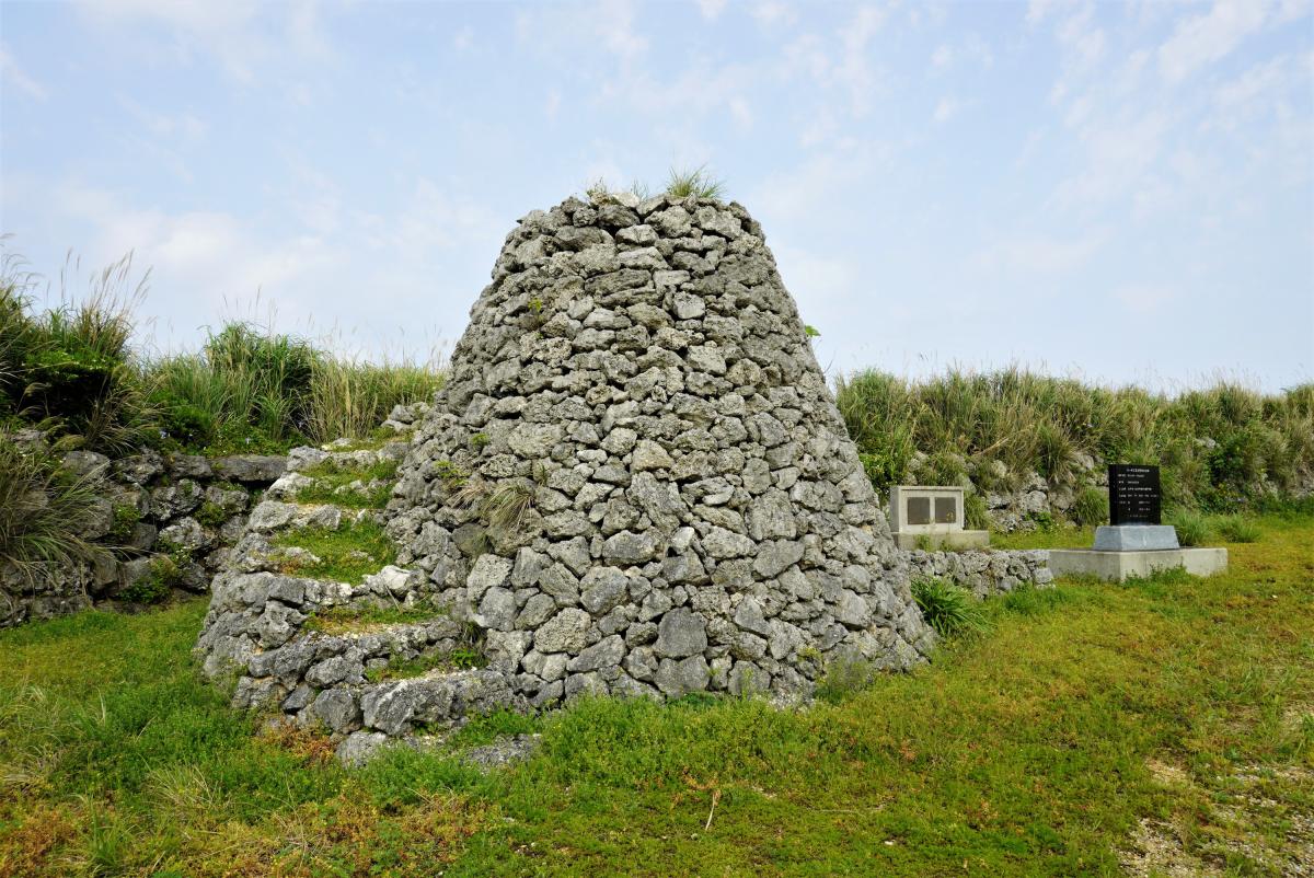 緑の草地の中央に、岩を積み上げて円錐形に造られた階段付きの石積みの見張り台と、右奥には石碑が見える写真