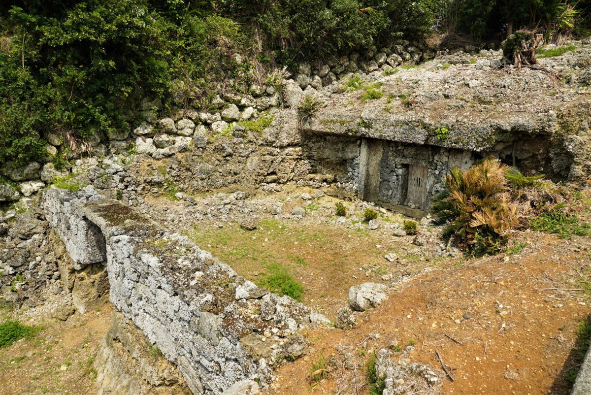緑の木々に囲まれた敷地の一角に、岩の石垣が築かれ、その中に扉がついた大きな石造りの納骨堂がある写真
