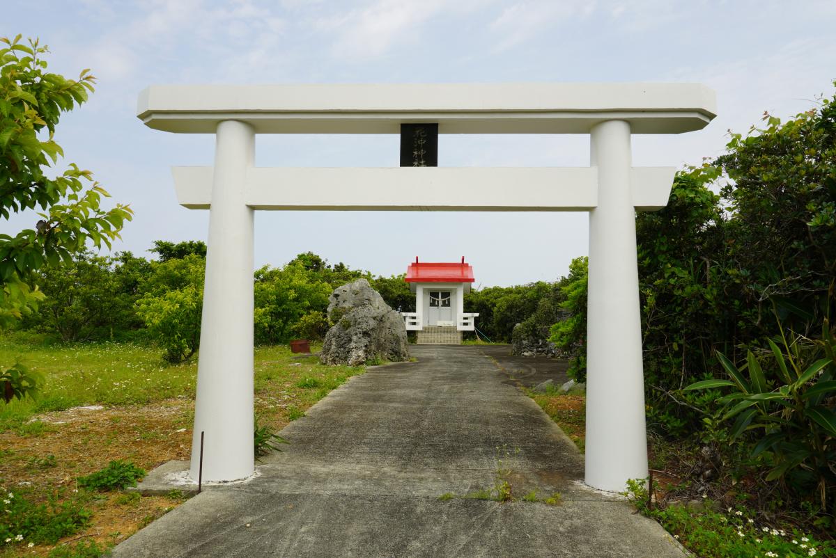 緑の木々に囲まれた敷地内に、白い鳥居が立ち、鳥居の奥に続く参道の先には大きな岩があり、さらにその奥には朱色の屋根の小さな祠が見える写真