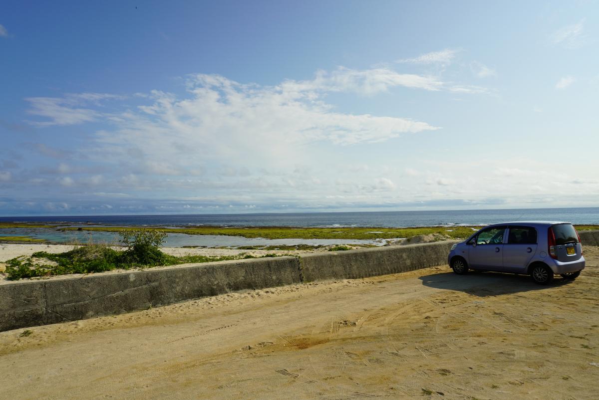 青い海と空、白い雲が広がる海岸線の手前にある砂利の広場に、レンタカーの軽自動車が駐車されている写真