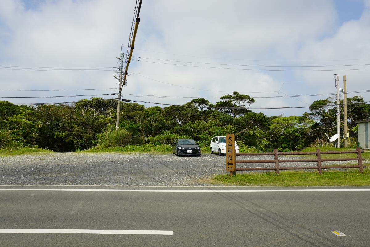 森の緑を背景にした砂利敷きの駐車場があり、黒と白の乗用車が駐車されており、右側には「大山神社入口」と書かれた木製の道標と柵が立っている写真