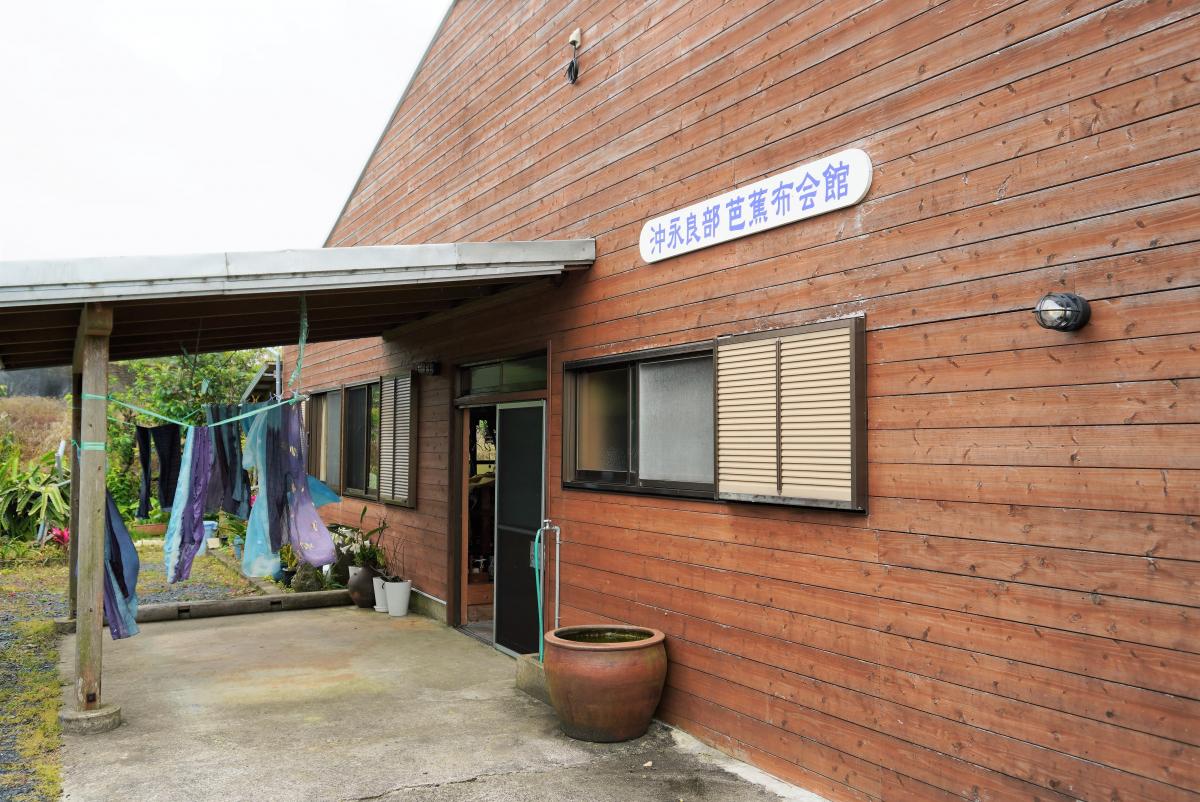 茶色の木目調の壁の建物と、左側にある屋根付きの入り口の付近に干されたカラフルな布、建物の壁には「沖永良部芭蕉布会館」と書かれた横長の看板が取り付けられている沖永良部芭蕉布会館の外観写真