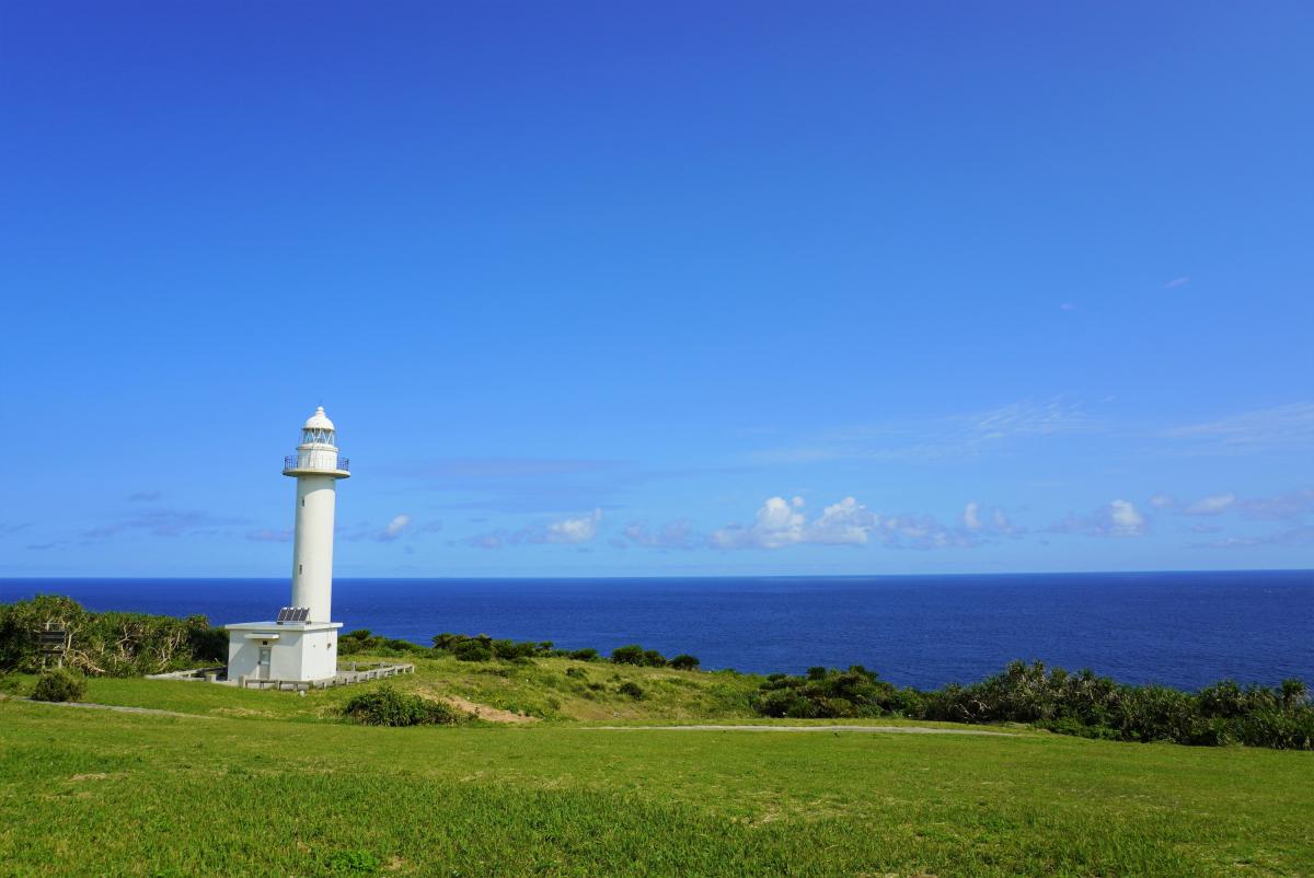 青い空と濃い青い海が広がる崖の上の緑の草地に、白く細長い灯台が立っている写真