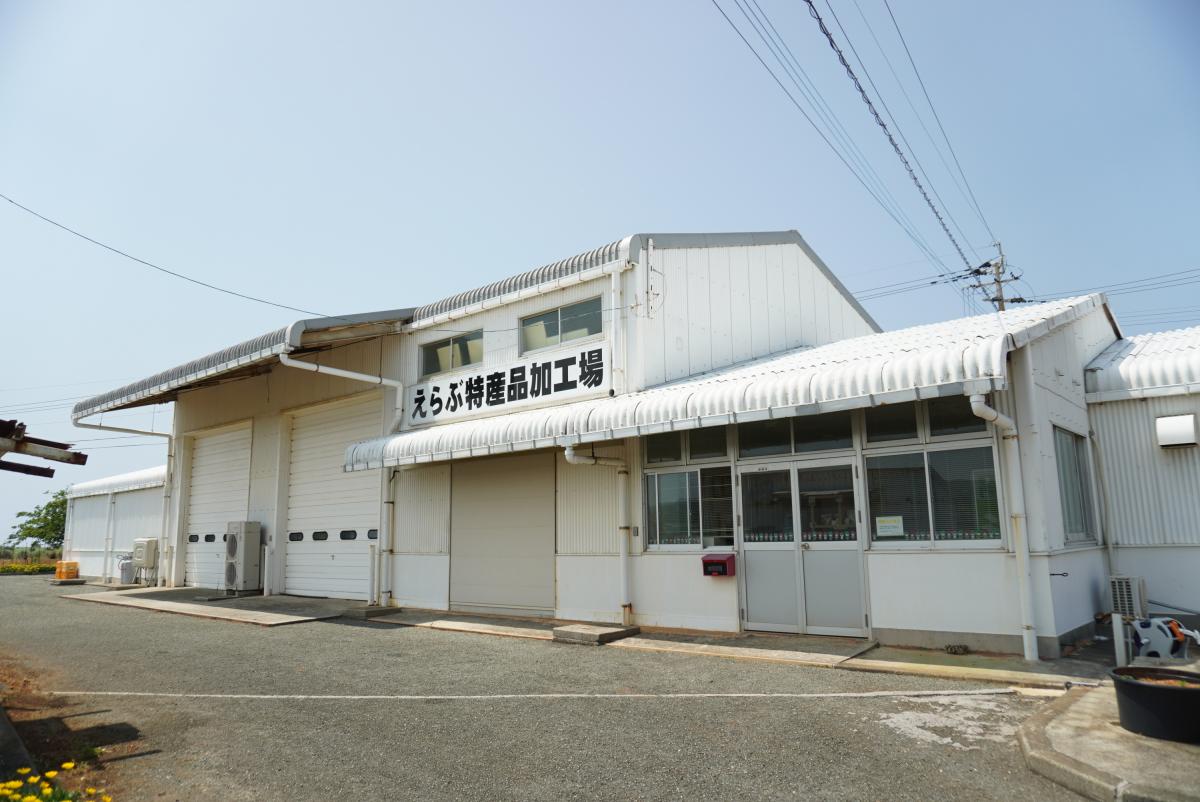 白い波板屋根とシャッターが特徴の平屋の工場兼事務所のような建物が写っており、中央上部に「えらぶ特産品加工場」と黒文字で書かれた看板があるえらぶ特産品加工場の外観の写真