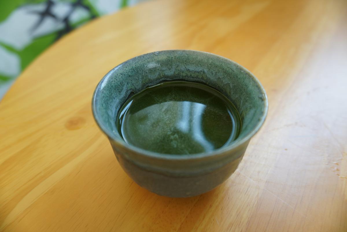 明るい木製のテーブルの上に置かれた、緑色の渋い色合いの陶器の湯呑みにシマ桑茶が注がれている写真