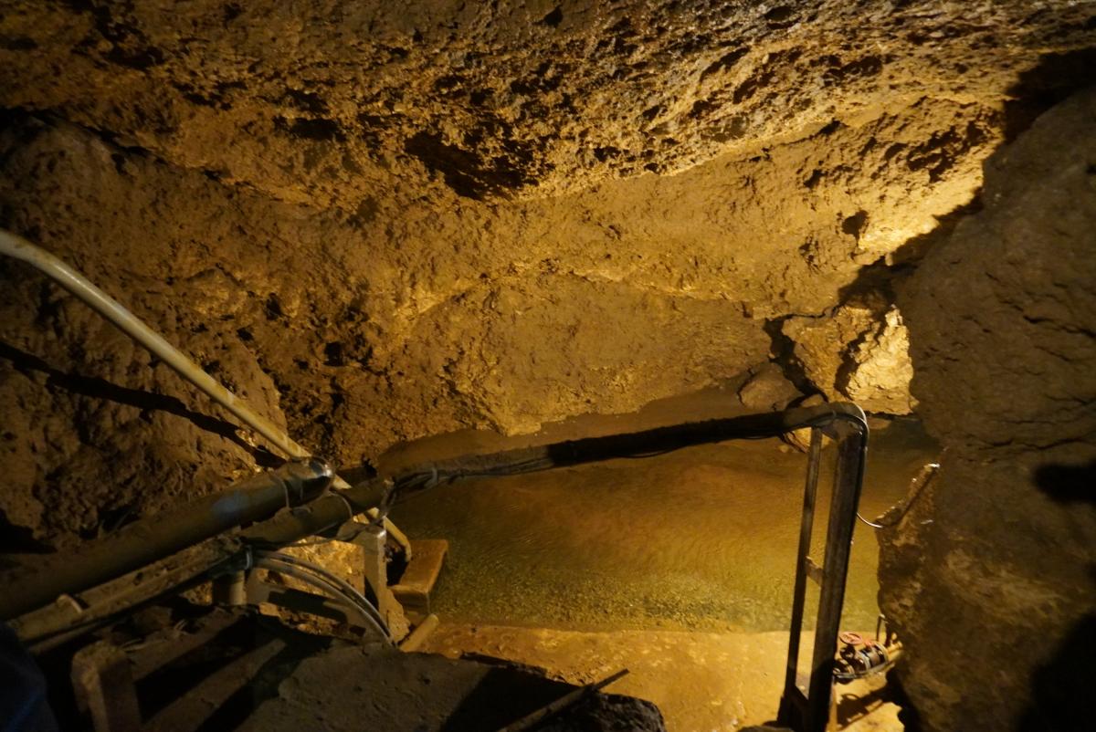茶色くごつごつした岩肌に囲まれた洞窟の内部が写っており、中央の水たまりの上には、金属製のパイプや手すりらしきものが設置されている写真