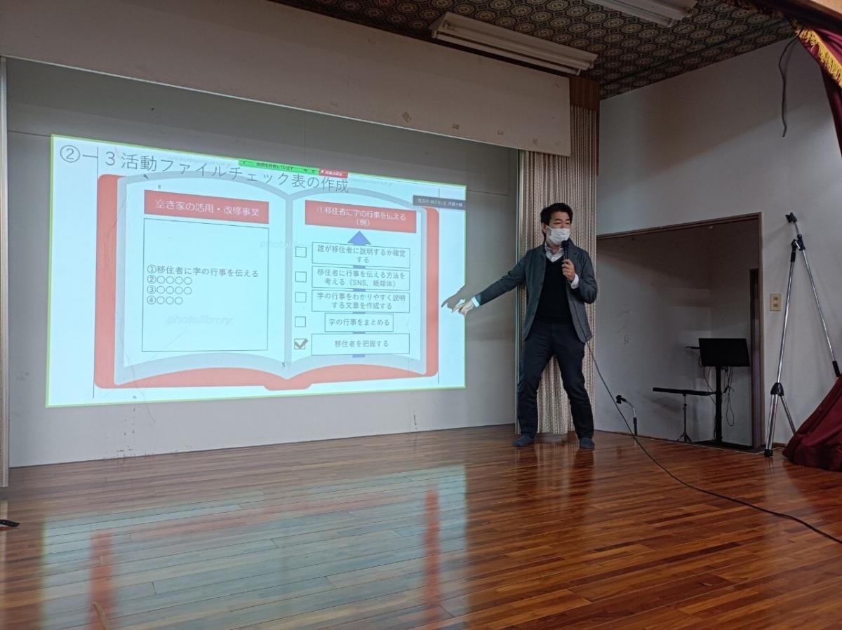 室内の舞台で、スーツ姿の人物がマイクを持ち、スクリーンに投影された赤枠の本のイラスト風スライドを指していて、スライドには「活動ファイルチェック表の作成」や複数のチェック項目が表示されている写真。