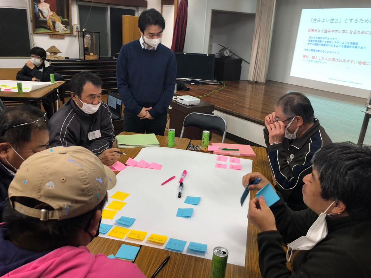 室内のテーブルで複数の人が集まり、中央の大きな白紙を囲んで青や黄色、ピンクの付箋を並べながら議論しており、背景のスクリーンには「住みよい住吉」とするための文章が投影されている写真。