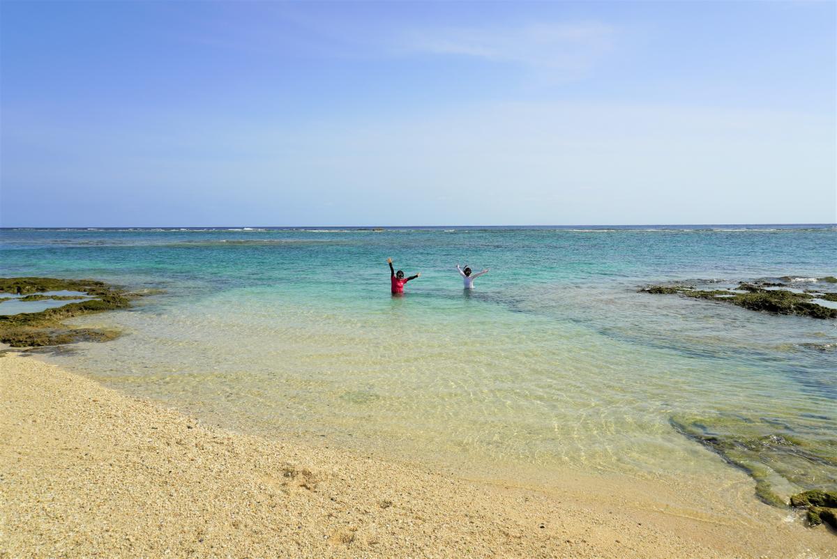 快晴の日の穏やかなビーチで、透明度の高い青い海の中に立っている二人が両手を上げている写真