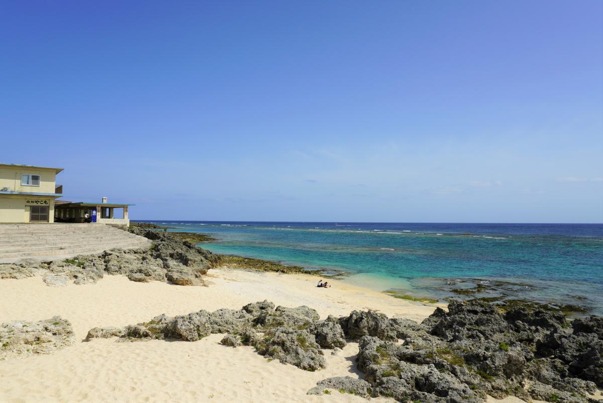 岩場と白い砂浜が広がる穏やかなビーチの奥に、ターコイズブルーの海と水平線が広がり、左奥には石階段が広がる写真
