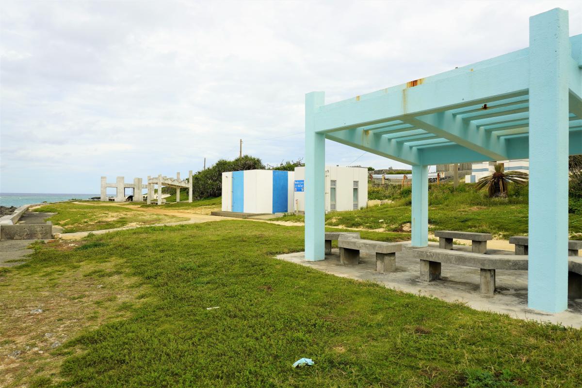 海辺の芝生の上に、水色の柱と梁の構造が特徴的な建物にベンチが置かれた休憩所があり、その奥には白と青に塗られた建物がある写真