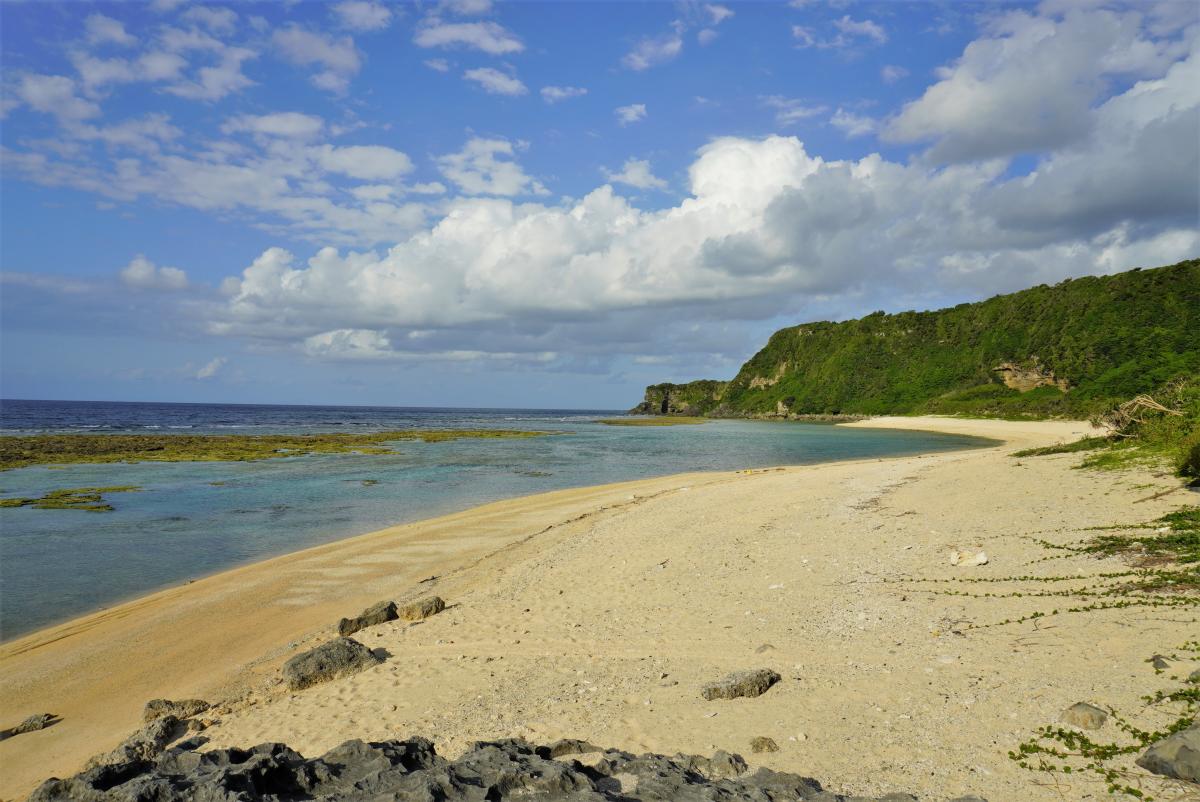 海と白い砂浜が広がり、右手には緑豊かな山がそびえ立ち、空には白い雲が浮かぶ沖泊海浜公園の写真