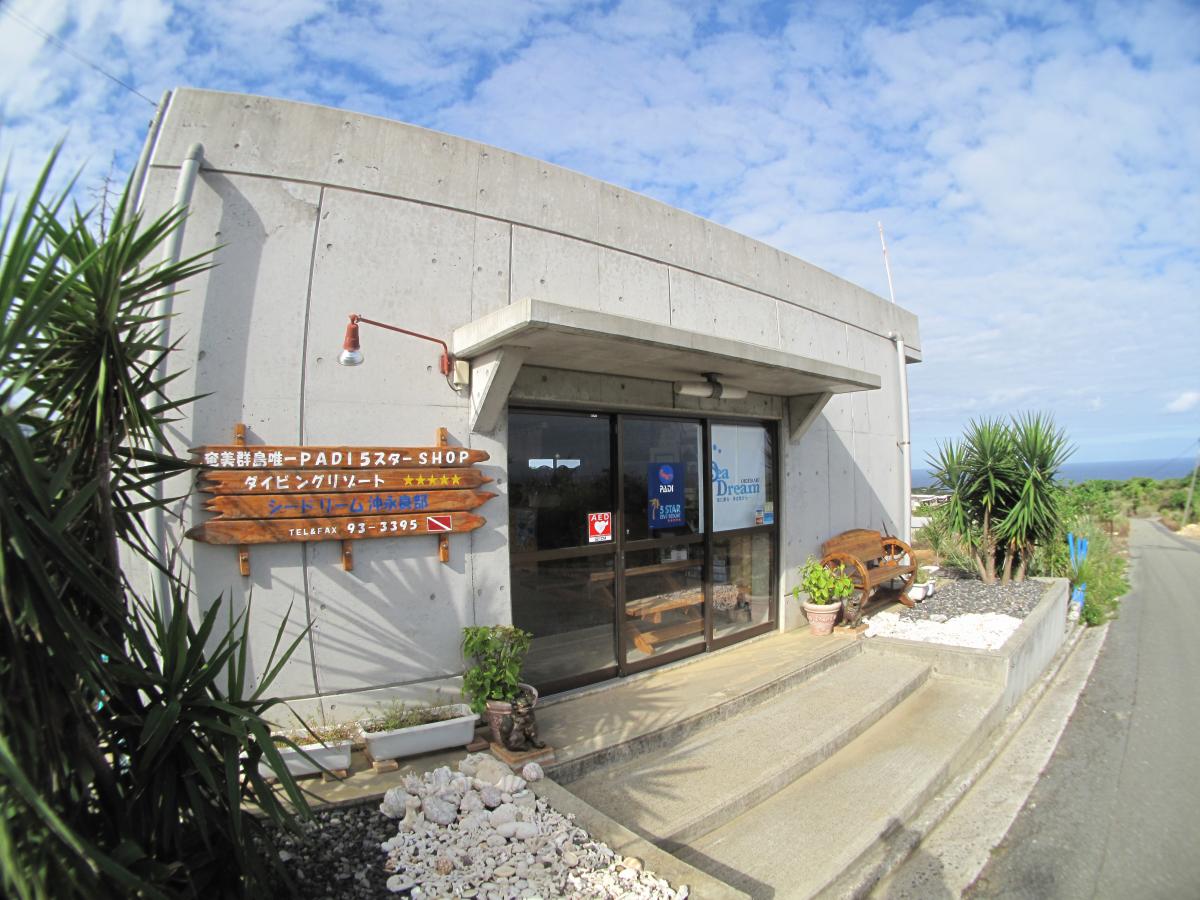 コンクリート造りの建物で、入口左には木製の看板が設置され両側には植木が植えられているダイビングの店シードリーム沖永良部の写真