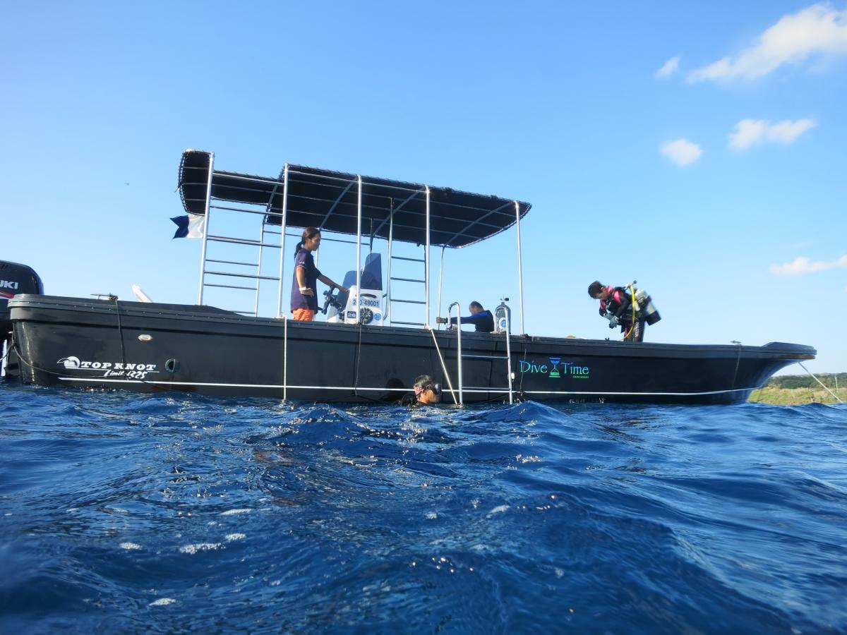 青い海と青空を背景に、ダイバーを乗せた黒いダイビングボートが浮かび、ダイビング器材を装着した人が船尾で準備し、水面には人が潜っている写真