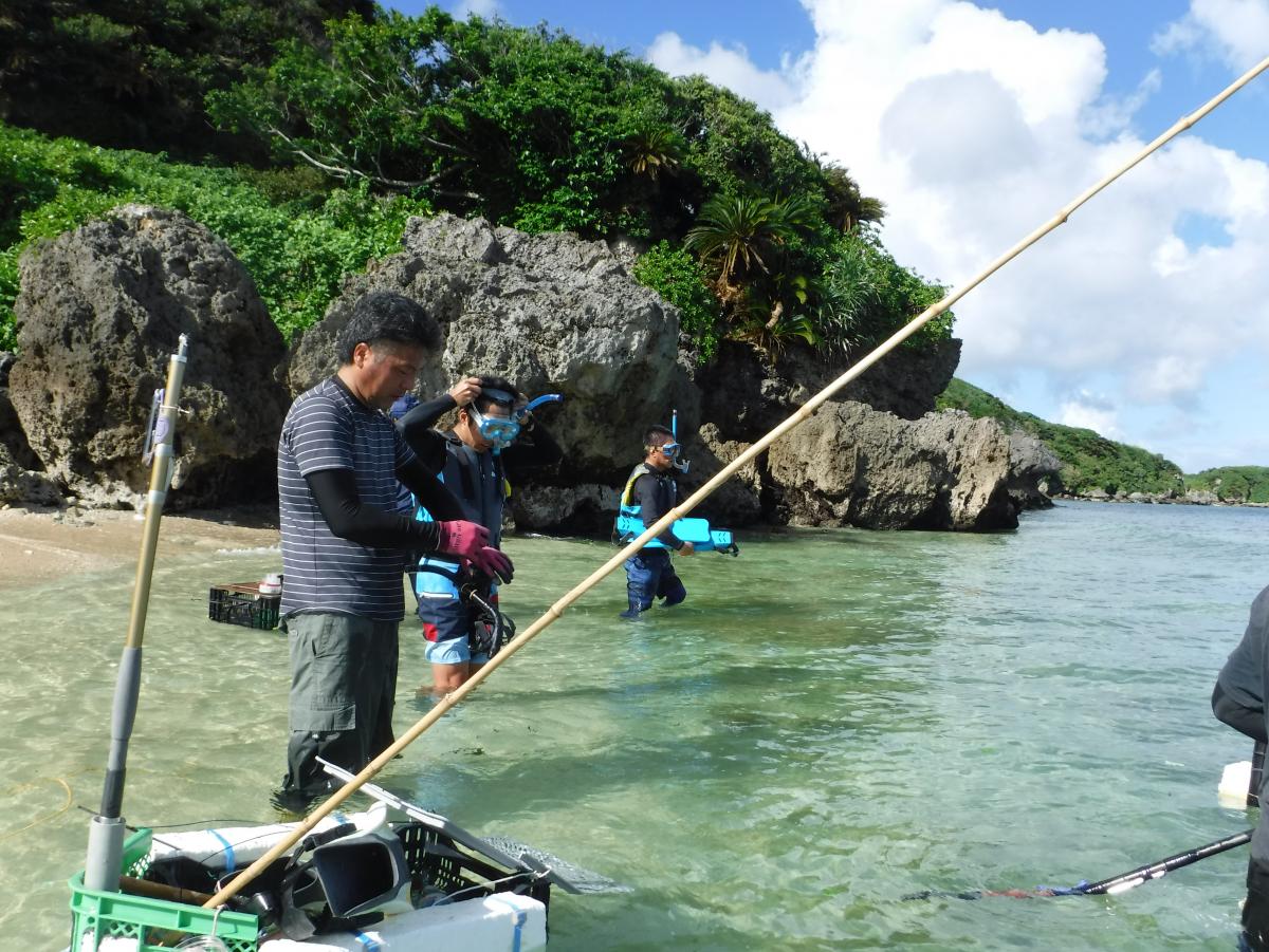 沖縄の海岸で、ライフジャケットを着てシュノーケルを装着した二人の男性と、水中に立つ男性が、魚釣りをしている様子の写真