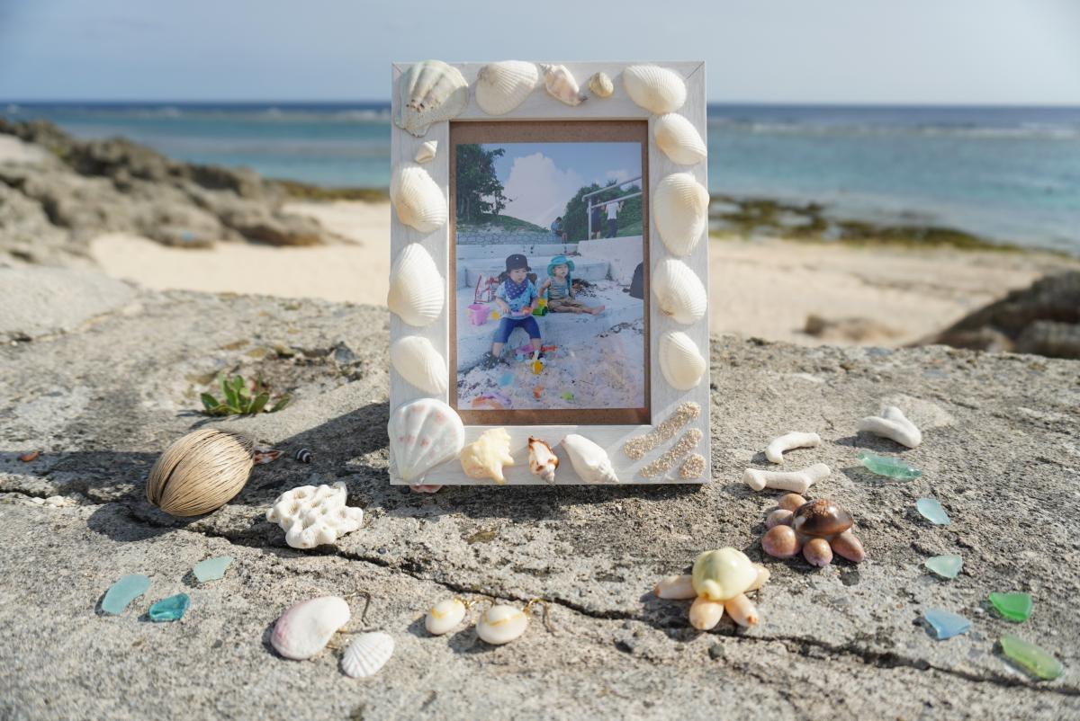 砂浜を背景にしたごつごつした岩の上に、貝殻やシーグラス、ビーチで拾ったと思われる小物が飾られ、その中央に貝殻で装飾されたフォトフレームがあり、中には砂場で遊ぶ二人の子どもの写真が収められている写真