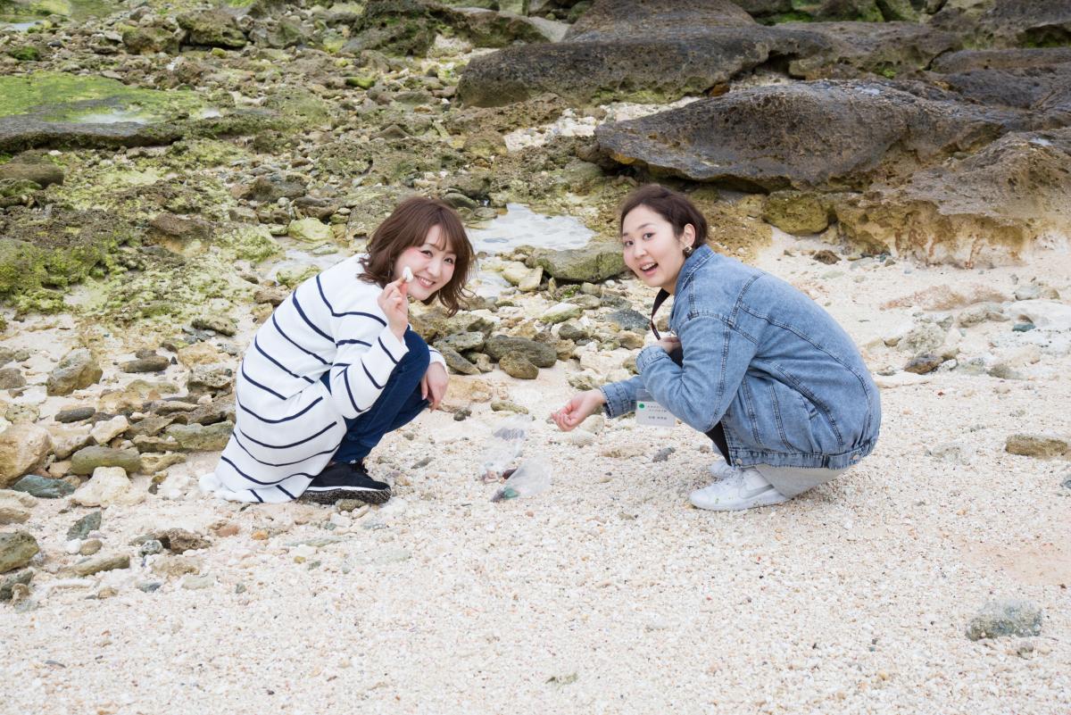 ごつごつした岩場に近い白い砂浜で、ストライプのトップスを着た女性とデニムジャケットを着た女性の二人が笑顔でしゃがみ込み、貝殻を集めている楽しそうな様子の写真