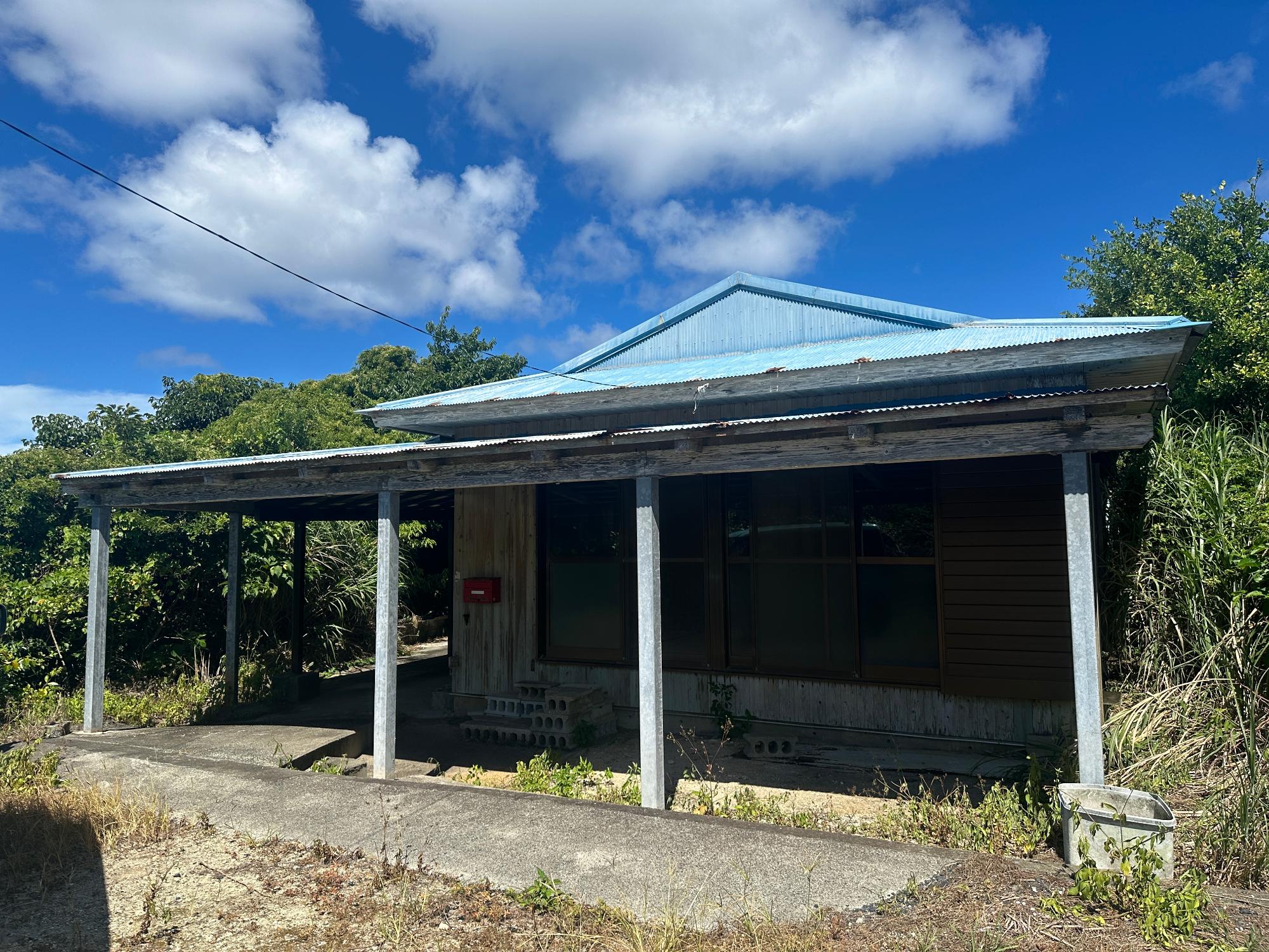 青いトタン屋根の木造平屋建ての建物が、建物正面全体に波板の庇が張り出し、その下を支える複数の細い金属柱と、深い緑の木々を背景に写る建物の外観写真