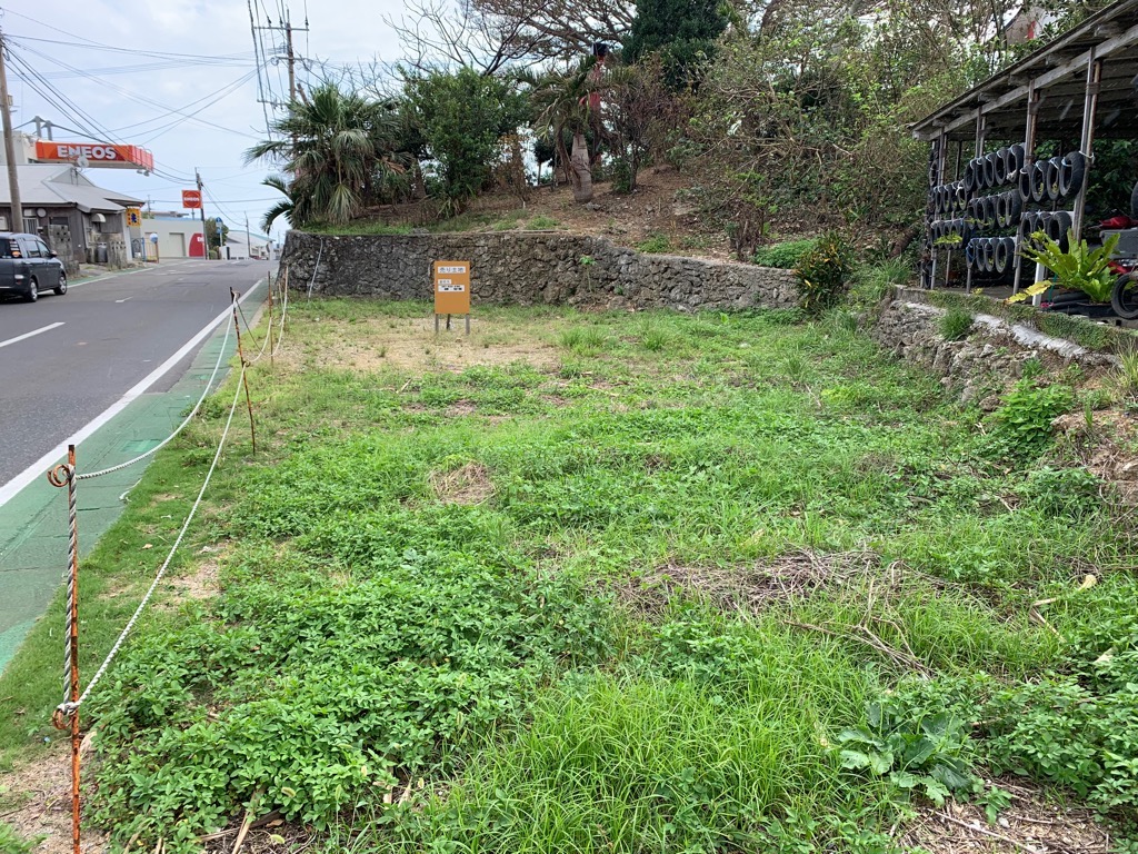 草が生い茂る空き地で、左にはアスファルトの道路と縁石、中央には「売り土地」の看板と石積みの擁壁、右奥にはタイヤなどが積まれた屋根付きの小屋が見える写真