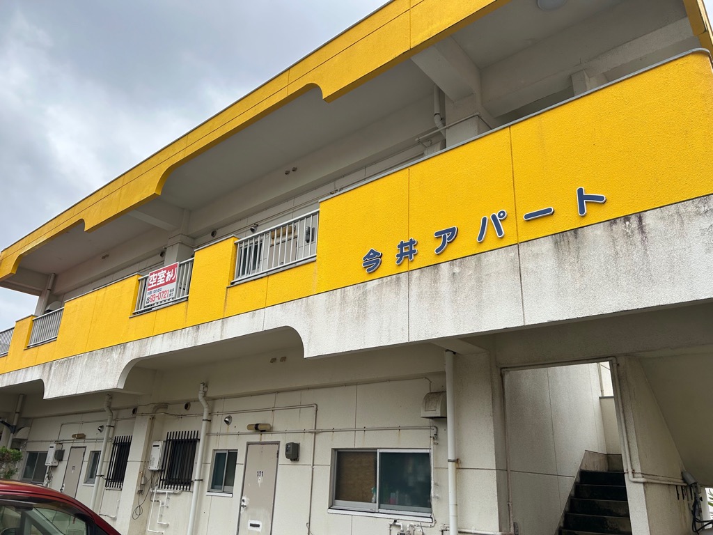 黄色のアプローチが目を引く2階建てのアパートで、建物の正面には「今井アパート」の文字が書かれ、1階の玄関や窓の周囲が白いアパートの外観写真
