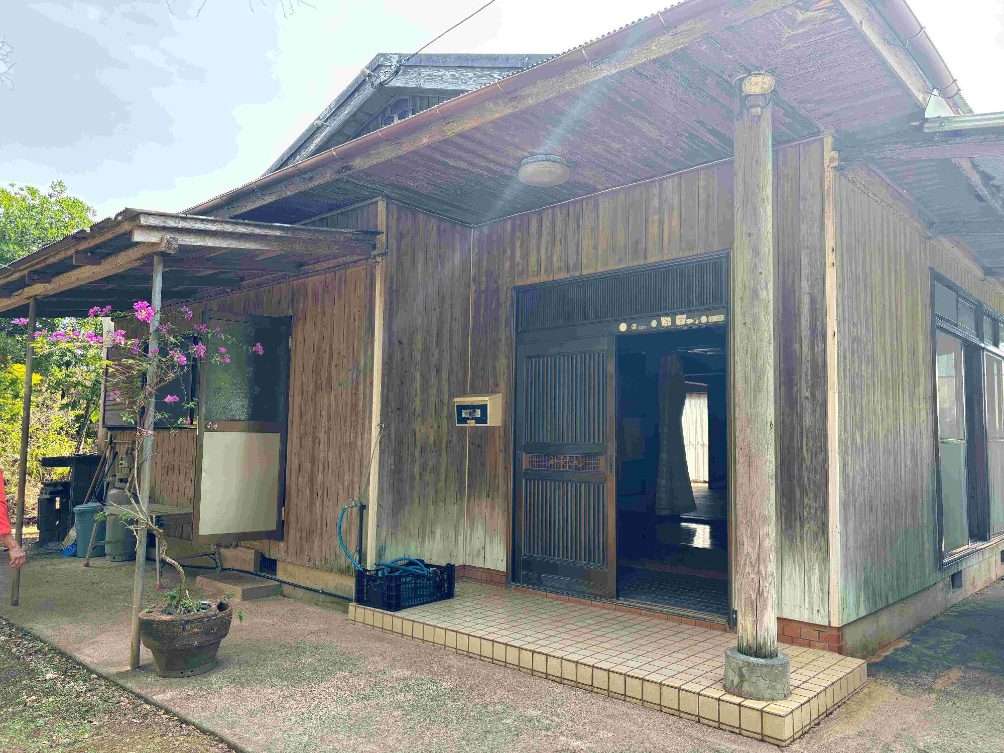 木造の一軒家で、正面の玄関は格子戸で開いており、手前にトタンの庇の下には植木鉢がある物件の外観写真
