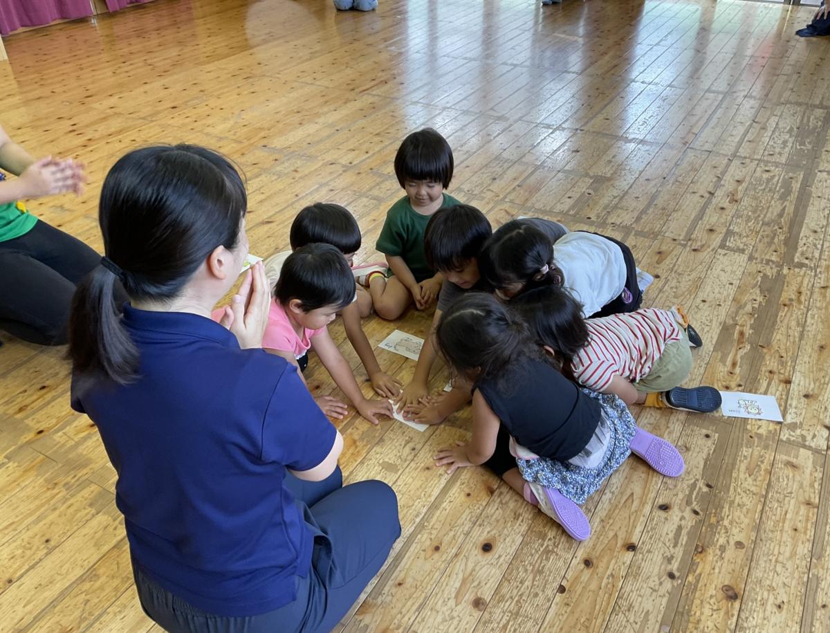 フローリングの室内で、園児たちが円になって座り、床に広げられたカードを取り合って遊んでいるのをポロシャツ姿の女性の先生が見ている写真