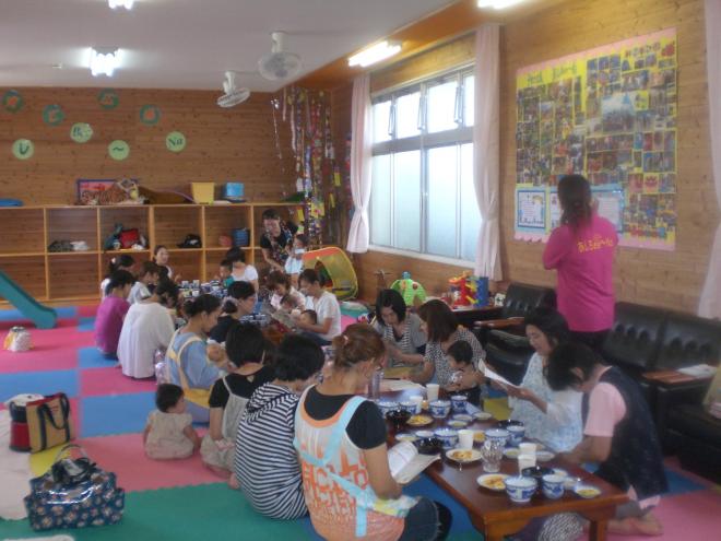 カラフルなマットが敷かれた室内で複数の大人と子どもがテーブルを囲み、交流をしている離乳食教室の様子の写真。