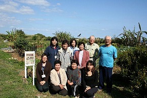 野菜畑の中で立つ男性や女性など総勢10名ほどの集団が、笑顔でカメラに向かって立ったりしゃがんだりしている集合写真