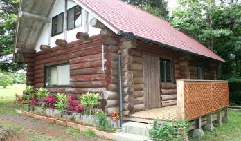 木々に囲まれた敷地に、赤い三角屋根と白い窓枠を持つロッジが建ち、外壁は丸太を積み重ねたデザインで、玄関前には木製の手すり付きデッキと花壇が整備された写真