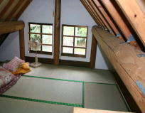 三角屋根の下にあるロフトスペースで、畳敷きの床に枕と布団が置かれ、木製の梁が見える天井と二つの窓から緑の景色が見える、シンプルで落ち着いた寝室の写真