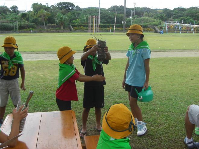 黄色い帽子をかぶり緑のスカーフを巻いた数人の子供たちが、緑の芝生が広がる広場で、ペットボトルでろ過装置を作り実験している様子の写真