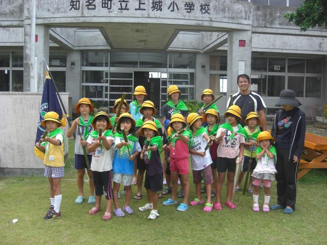 黄色い帽子と緑のスカーフを着用した子供たちが、「知名町立上城小学校」と書かれた校舎の玄関前にある芝生の上で、竹で出来た水鉄砲を手に持って旗と共に記念撮影をしている集合写真