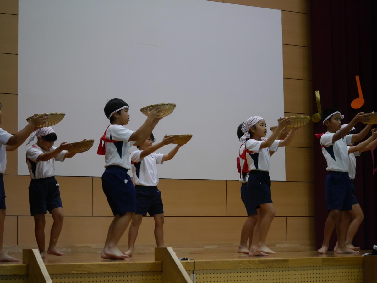 体育館のステージ上で、体操服姿に鉢巻を巻き、稲刈りの道具である脱穀用の「箕（み）」のような籠を頭上に掲げながら、素足で踊る複数人の小学生たちの写真