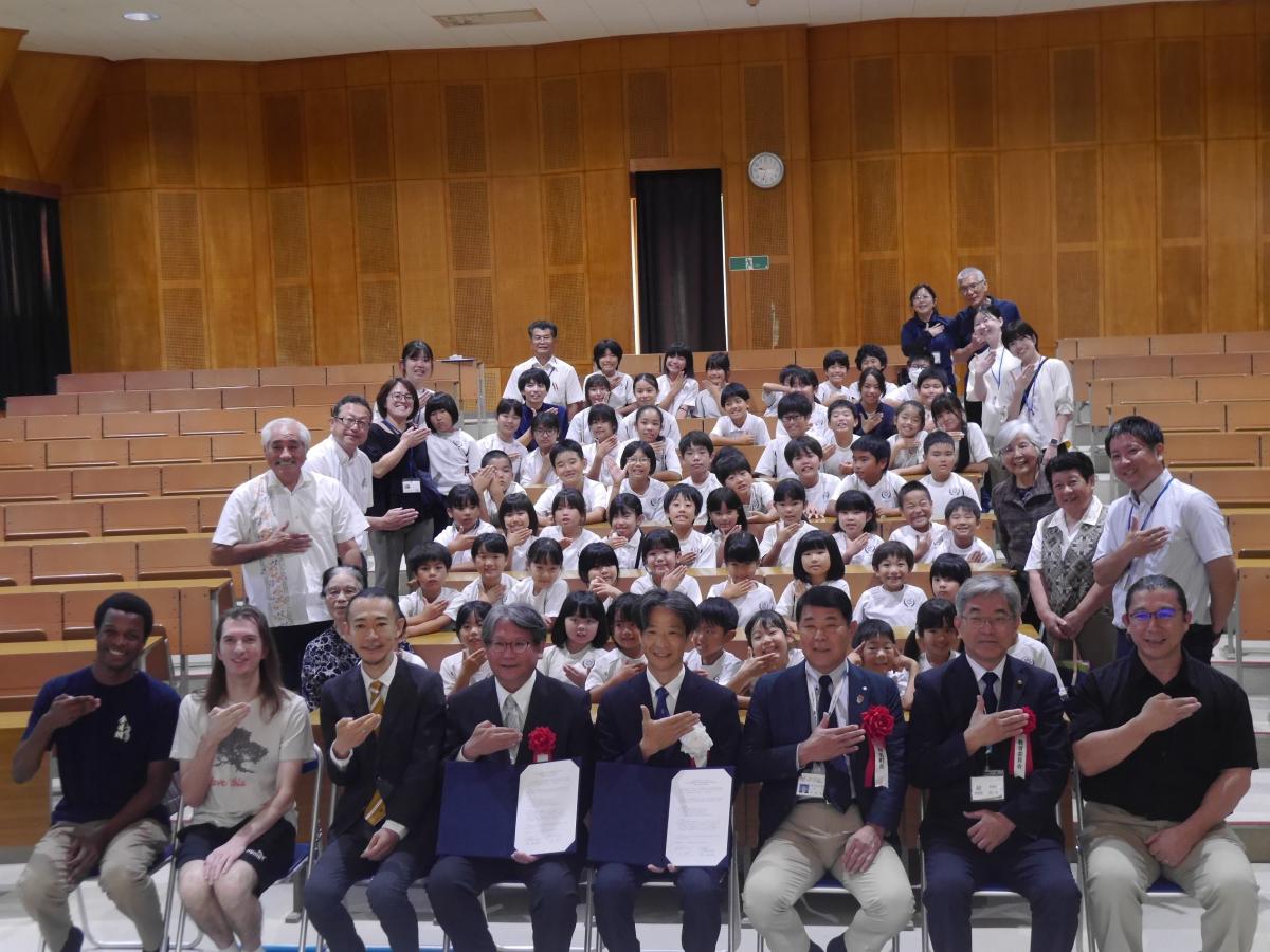 ひな壇式の座席が特徴的な講堂で、大人の関係者と多数の児童が一堂に会し、最前列の関係者の方々が協定書を抱え、全員が右手を胸に当てて写っている記念写真