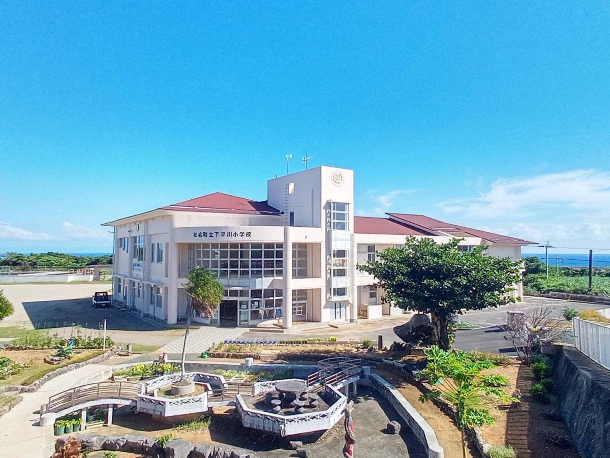 海岸沿いの高台に建つ淡いピンク色の「知名町立下平川小学校」の校舎が中央にあり、手前には橋やテーブルが配置された庭園のようなエリアが見え、建物の奥には青い海が広がる下平川小学校の外観写真