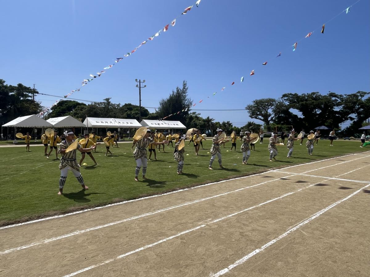 芝生が広がる運動場で、伝統的な衣装を身に着けた大勢の児童たちが踊っている様子の写真