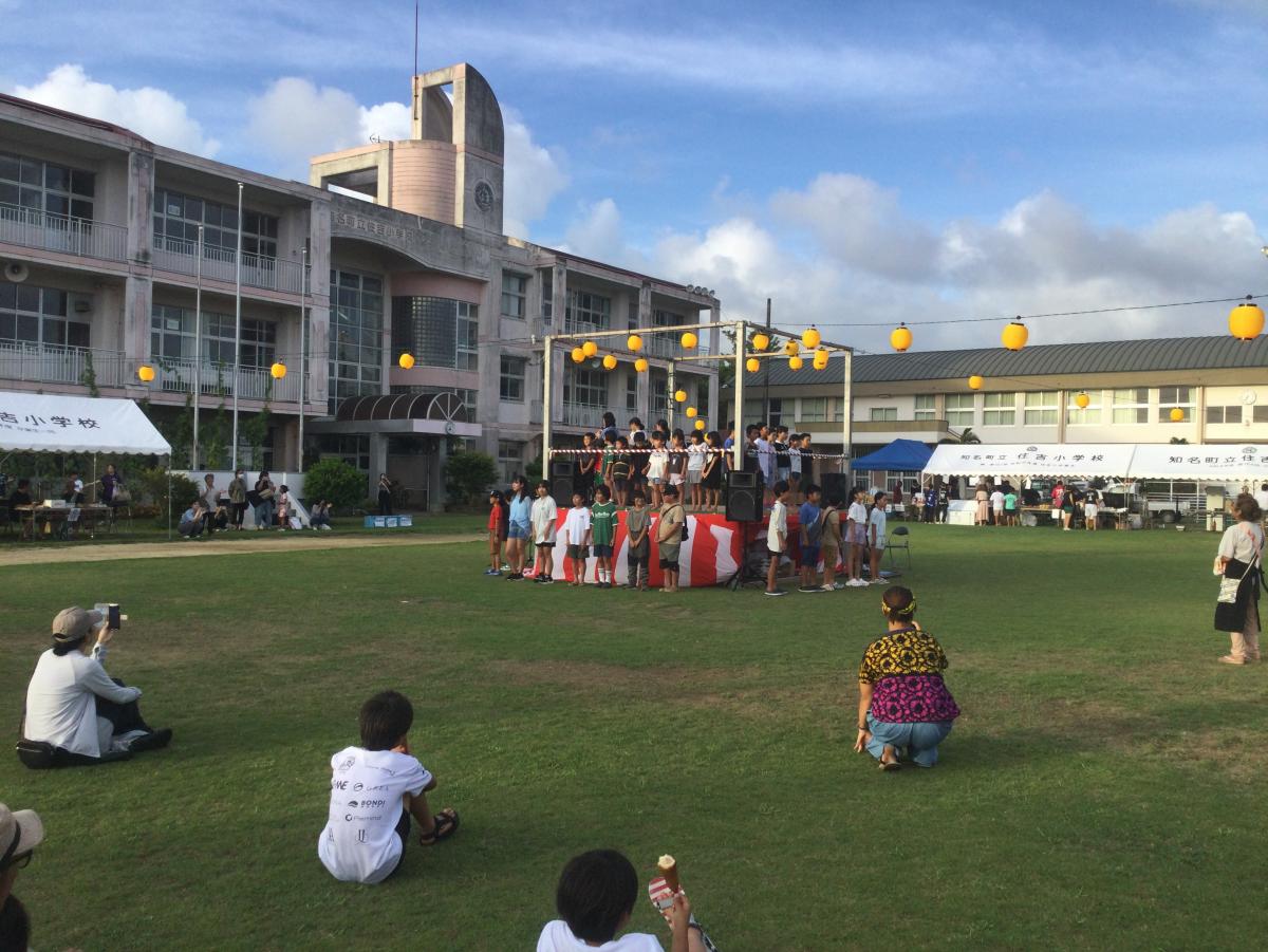 校庭の中央に組まれたやぐらの上に大人や児童たちが立っており、仮設ステージを取り囲むように多くの生徒や人が集まっている夕方の祭りの様子の写真