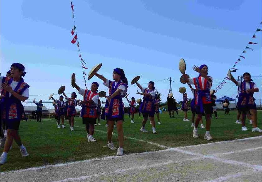 鉢巻と紫と白の衣装を着た児童たちが、笠を手に持ち一斉に踊っている運動会の写真
