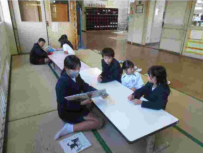 畳敷きの部屋で、黒いブレザーを着た女子生徒が、中央の白い長机の奥で、左右に座る2人の児童に絵本を見せながら読み聞かせをしている写真