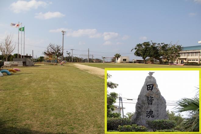 広大な芝生と土のグラウンドが広がる学校の校庭の風景と左奥には三本の旗竿に掲げられた国旗と校旗が見え、中央右下に拡大された、「田皆魂」と縦書きで刻まれた特徴的な形の巨大な石碑が写っている写真