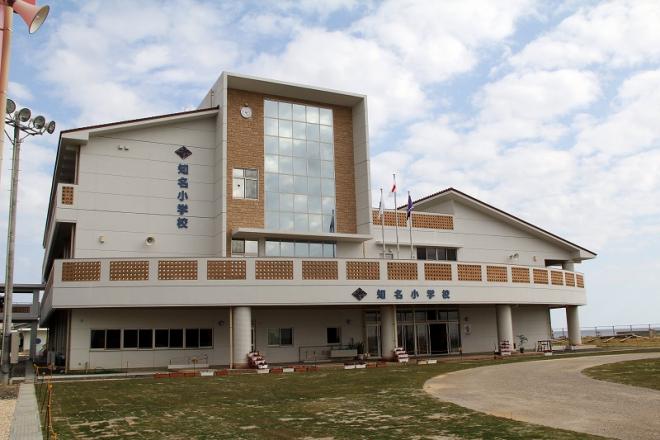 正面中央に大きなガラス窓と縦に「知名小学校」と書かれた文字が配置され、茶色のタイルと白い壁の近代的な二階建て校舎の知名小学校の外観写真