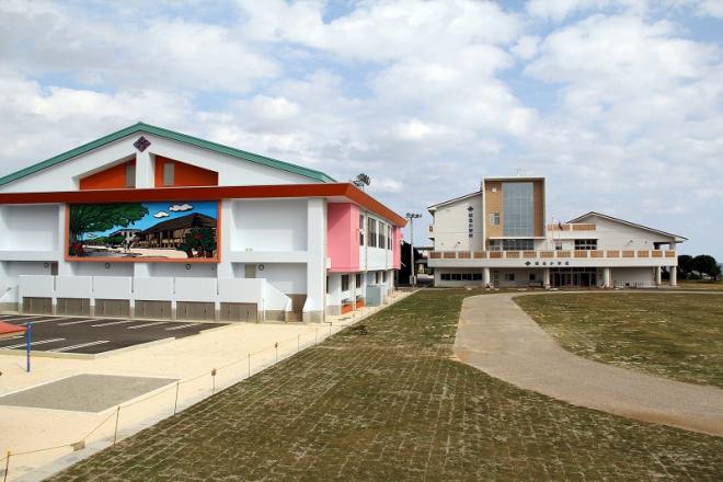 地域風景の大きな壁画が描かれ、ピンクのアクセントが施された体育館が左側にあり、校庭の億に近代的な校舎が見える学校の全景写真