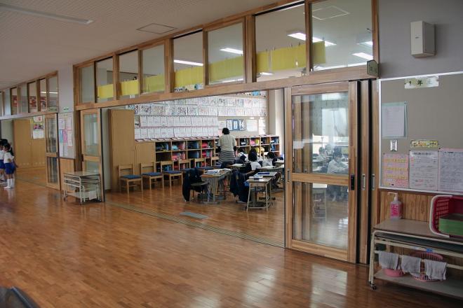 明るい木目調の床と柱に大きなガラスの引き戸が並んだ廊下から、児童と先生が活動している教室の様子を写した屋内写真