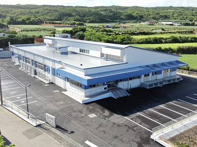 青い帯状のデザインが施された2階建ての建物が写っており、正面に屋根付きの出入口や搬入口が見え、周囲には舗装された敷地とその奥に緑の畑や低い丘が広がっている給食センターの外観写真